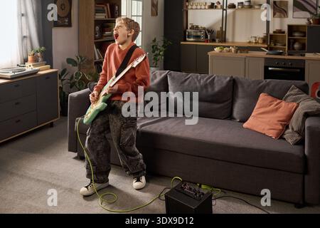 Bambino caucasico in piedi in salotto che suona la chitarra elettrica e canta con la bocca aperta, tenendo lo strumento con entrambe le mani, amplificatore e cavo sul pavimento, cucina sullo sfondo Foto Stock