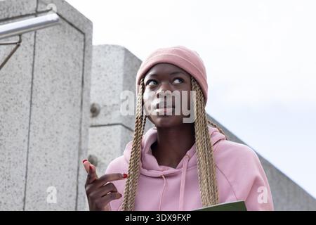 Donna afroamericana in berretto rosa e felpa con cappuccio da un muro di cemento che tiene la cartella verde e punta Foto Stock