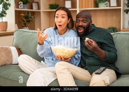 Una felice coppia multirazziale si siede a stretto contatto su un divano verde, guardando la tv e mangiando popcorn. Foto Stock