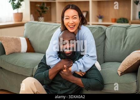 Allegra coppia interrazziale condivide un gioioso momento di risate mentre si rilassa a casa. Stanno abbracciando e guardando la telecamera. Foto Stock