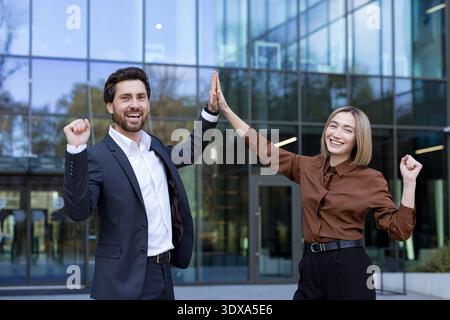 Due allegri uomini d'affari, un uomo e una donna, in piedi fuori da un edificio di uffici, festeggiano i successi e una partnership di successo con un alto gesto di cinque Foto Stock