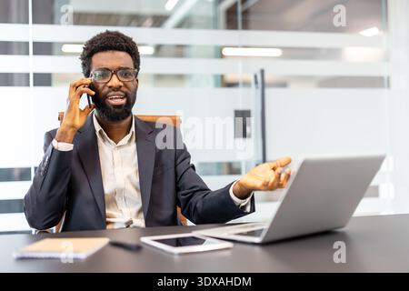 Uomo professionale che ha un'importante conversazione telefonica alla propria scrivania in un ufficio moderno, gestando durante la comunicazione dei concetti aziendali, utilizzando il proprio notebook e tablet Foto Stock