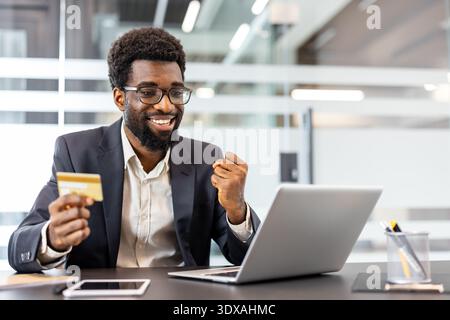 Uomo d'affari afro-americano sorridente allegramente e pugilato, tenendo in mano una carta di credito mentre effettua un acquisto online o completa una transazione su un computer portatile alla scrivania di un ufficio Foto Stock