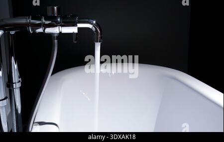 Primo piano di un moderno rubinetto cromato con un flusso costante di acqua dolce che fluisce in una vasca da bagno in ceramica bianca. L'immagine mostra un de minimalista Foto Stock