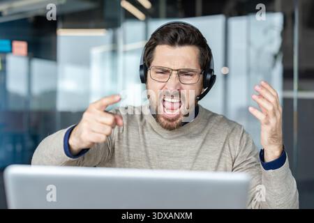 Uomo che indossa cuffie e occhiali, esprime rabbia e frustrazione estrema, punta il dito con una bocca aperta mentre lavora su un notebook in un ufficio moderno Foto Stock