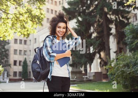 Una persona sorridente sta all'esterno con zaino e cartella mentre si gode una giornata di sole al campus Foto Stock