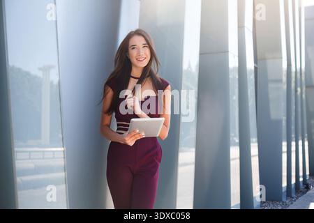 Una giovane donna usa il tablet mentre si appoggia all'edificio moderno durante il giorno in un ambiente urbano Foto Stock