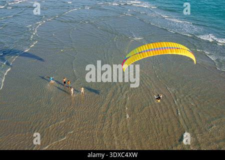 Vista aerea di un parapendio che si innalza sopra una famiglia che si tuffa attraverso le acque poco profonde baciate dal sole, dove il mare turchese incontra la spiaggia sabbiosa Jupit Foto Stock
