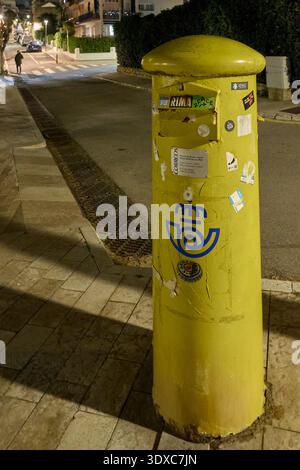 Sitges, Spagna - 02 marzo 2026: Cassetta postale gialla di Correos su una strada di notte con adesivi e texture urbana in un quartiere residenziale. Foto Stock
