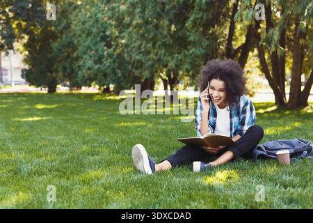 La ragazza siede su erba e parla al telefono mentre legge un libro in un parco Foto Stock