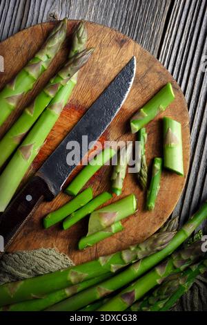 Gli asparagi verdi impacchettati sono legati con un coltello posto su un tagliere di legno rotondo e verdure biologiche fresche. Disposizione piatta, vista dall'alto Foto Stock