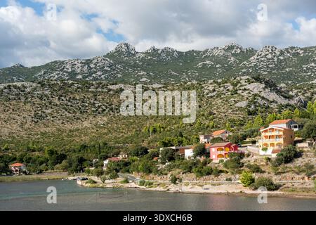 Piccolo villaggio sul mare croato con case colorate lungo la baia tranquilla, la strada costiera e la spettacolare catena montuosa carsica ricoperta da una foresta verde alle spalle. Con Foto Stock