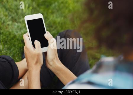 Persona seduta su erba con in mano uno smartphone mentre si gode un momento di tempo libero all'aperto Foto Stock