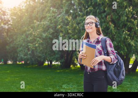 Una giovane donna ama ascoltare musica in un parco mentre tiene in mano libri e indossa le cuffie durante il giorno Foto Stock
