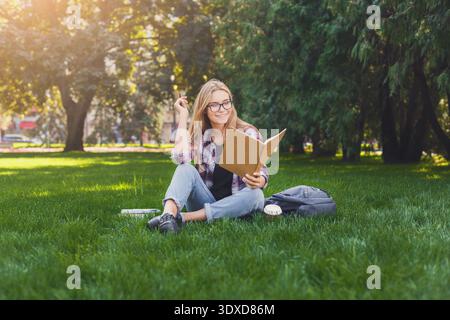 Una giovane donna siede sull'erba e ama leggere un libro in un parco durante la giornata di sole Foto Stock
