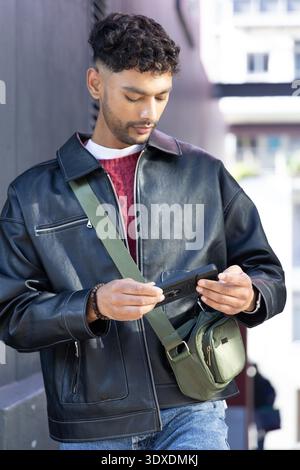 Uomo indiano appoggiato al muro scuro per strada, tenendo il telefono, indossando giacca nera e borsa verde Foto Stock