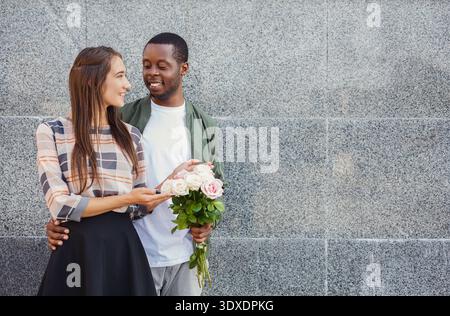 Coppia sorridi e condividi momenti mentre ti trovi in piedi contro la parete grigia tenendo i fiori Foto Stock