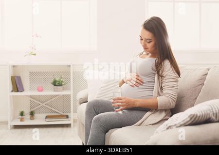 Donna incinta siede sul divano in una stanza accogliente con un bicchiere d'acqua in mano Foto Stock