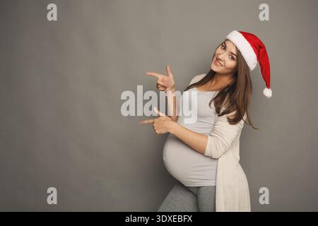 Donna incinta che indossa Santa Hat Poses and Points allo Space for Holiday Collage Foto Stock