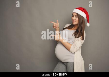 Una donna incinta che indossa un cappello di Babbo Natale posa e punti durante una sessione fotografica di festa Foto Stock