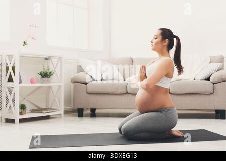Aspettarsi che la madre pratichi lo yoga a casa su un comodo tappetino nello spazio luminoso del soggiorno Foto Stock