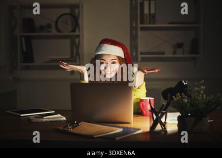 Una donna con capelli ricci rossi siede a una scrivania in un ufficio domestico con un cappello di Babbo Natale Foto Stock