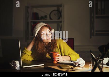 La donna di Santa Hat siede alla scrivania con laptop e tazza in un ambiente accogliente Foto Stock