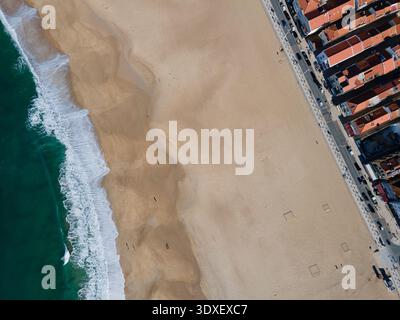 Vista aerea del punto d'incontro delle onde turchesi dell'oceano con la spiaggia sabbiosa, in contrasto con gli edifici dal tetto rosso, Nazaré, il quartiere di Leiria Foto Stock