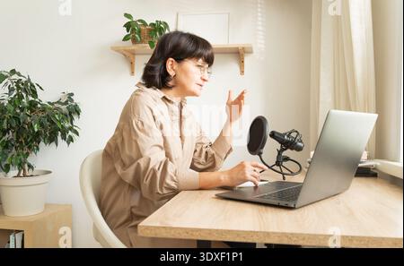 Podcasting donna caucasica matura dall'ufficio domestico con configurazione laptop e microfono Foto Stock