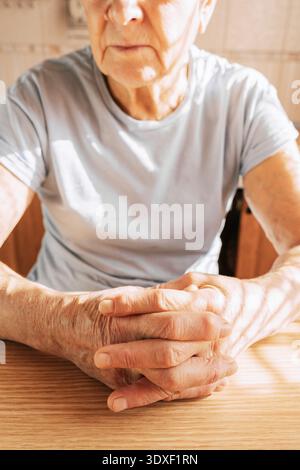 Una donna anziana siede a un tavolo in una stanza soleggiata, le mani piegate sul tavolo, primo piano delle mani Foto Stock