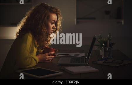 Donna che fissa il laptop con un caffè in mano durante le ore serali a Home Space Foto Stock
