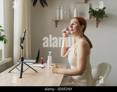 Donna che crea un vlog di bellezza che applica un massaggiatore facciale. Un beauty blogger registra i contenuti per i social media utilizzando uno smartphone Foto Stock