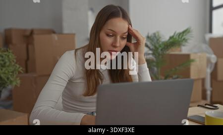 Giovane donna ispanica al portatile con la mano sul tempio, che poggia il gomito sul tavolo, circondata da scatole di cartone nell'edificio; transizione in movimento stressata. Foto Stock