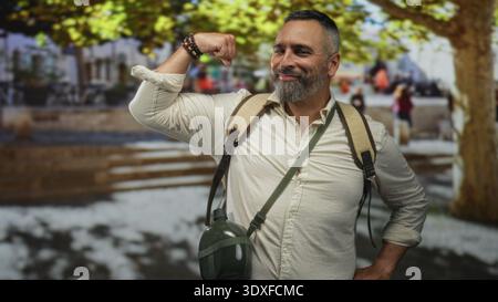 L'uomo ispanico di mezza età si flette bicipite, sorridendo, indossando zaino con mensa e braccialetti in Street plaza; avventura sicura. Foto Stock