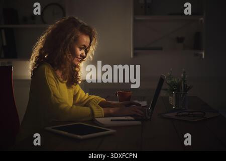 Donna che lavora a tarda notte con un laptop in un ambiente di ufficio domestico con luce calda e un'atmosfera accogliente Foto Stock