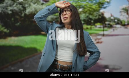 La donna protegge gli occhi con la mano indossando occhiali e blazer sul percorso illuminato dal sole del parco stradale guardando verso l'alto; curiosità attesa. Foto Stock