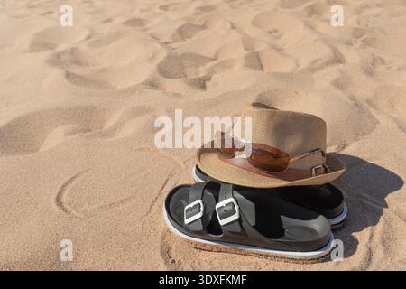 Primo piano di un cappello di paglia marrone, occhiali da sole e sandali neri adagiati su una spiaggia sabbiosa alla luce del sole. Tema delle vacanze Foto Stock