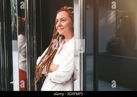 Una moderna donna d'affari si trova davanti al business center con le braccia incrociate. Ritratto di donna con dreadlock. Donna di mezza età con acconciatura insolita || modello rilasciato Foto Stock