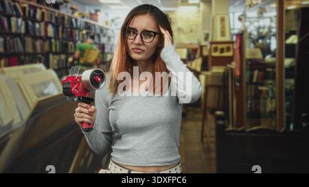 Donna che tiene in mano un dispenser di nastro e che afferra un rotolo di nastro da imballaggio, tocca il tempio e indossa gli occhiali in un corridoio della libreria di un edificio; frustrazione Foto Stock