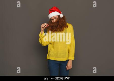 Donna con capelli ricci che indossa un cappello rosso Santa e un maglione giallo che tiene i capelli in un ambiente Studio Foto Stock