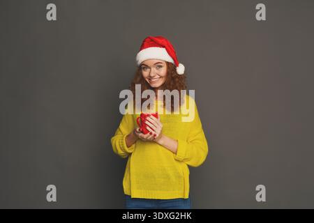 Donna sorridente in un cappello rosso che regge una tazza su sfondo grigio in inverno Foto Stock