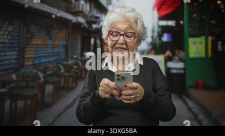Donna anziana nonna dai capelli bianchi che indossa gli occhiali che reggono lo smartphone, tocca lo schermo con i pollici in un bar di strada con sedie e graffiti; digita Foto Stock