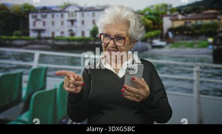 Donna anziana che punta il dito e tiene lo smartphone con una mano mentre tocca lo schermo, indossa occhiali colorati e un maglione accanto a una struttura sul lungomare Foto Stock