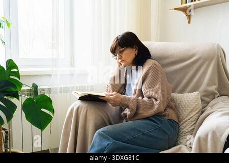Donna in occhiali che si rilassa in una poltrona vicino alla finestra, infilata in un libro, godendo del tempo libero a casa Foto Stock