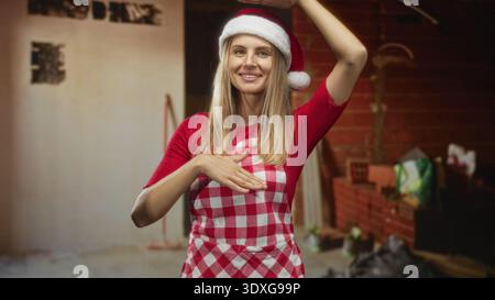 Donna con cappello di babbo natale e grembiule a scacchi, mano posta sul petto in una cucina di un edificio in mattoni; gioia per le vacanze. Foto Stock