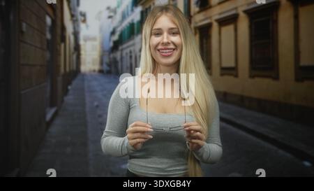 Donna che tiene gli occhiali da vista e sorride su una stretta strada acciottolata tra vecchi edifici, capelli lunghi visibili e scollatura; sicurezza. Foto Stock