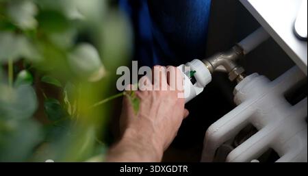 La persona regola la valvola sul radiatore di riscaldamento mentre si trova all'interno vicino a un impianto Foto Stock