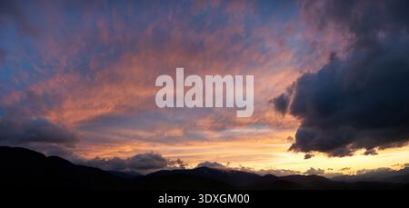 Spettacolare tramonto con nuvole scure e scoscese che proiettano ombre sul cielo. Horizon si illumina di calde tonalità arancio e rosa, contrastando con le silhouette delle montagne, creando un'atmosfera soffusa. Foto Stock
