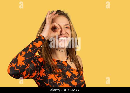 Giovane donna felice con dreadlocks e piercing sorridenti e che guarda attraverso il suo gesto di mano Foto Stock