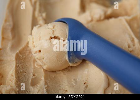 Una foto macro dettagliata con un cucchiaio per gelato blu che incide un cucchiaio omogeneo e uniforme da un lotto di cremoso gelato al burro di arachidi Foto Stock